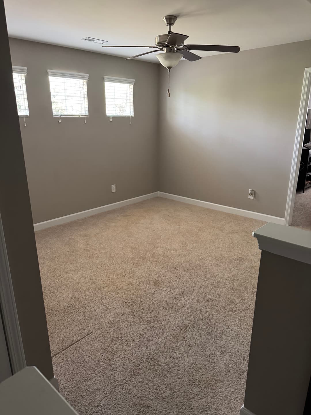 Empty room with beige carpet, fan, beige walls, and three windows letting in natural light.