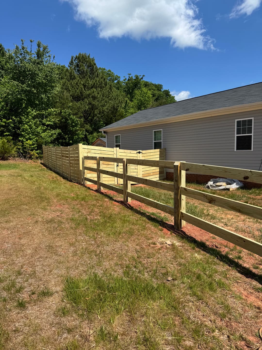 Wooden fence installation next to a house and green landscape under a blue sky.