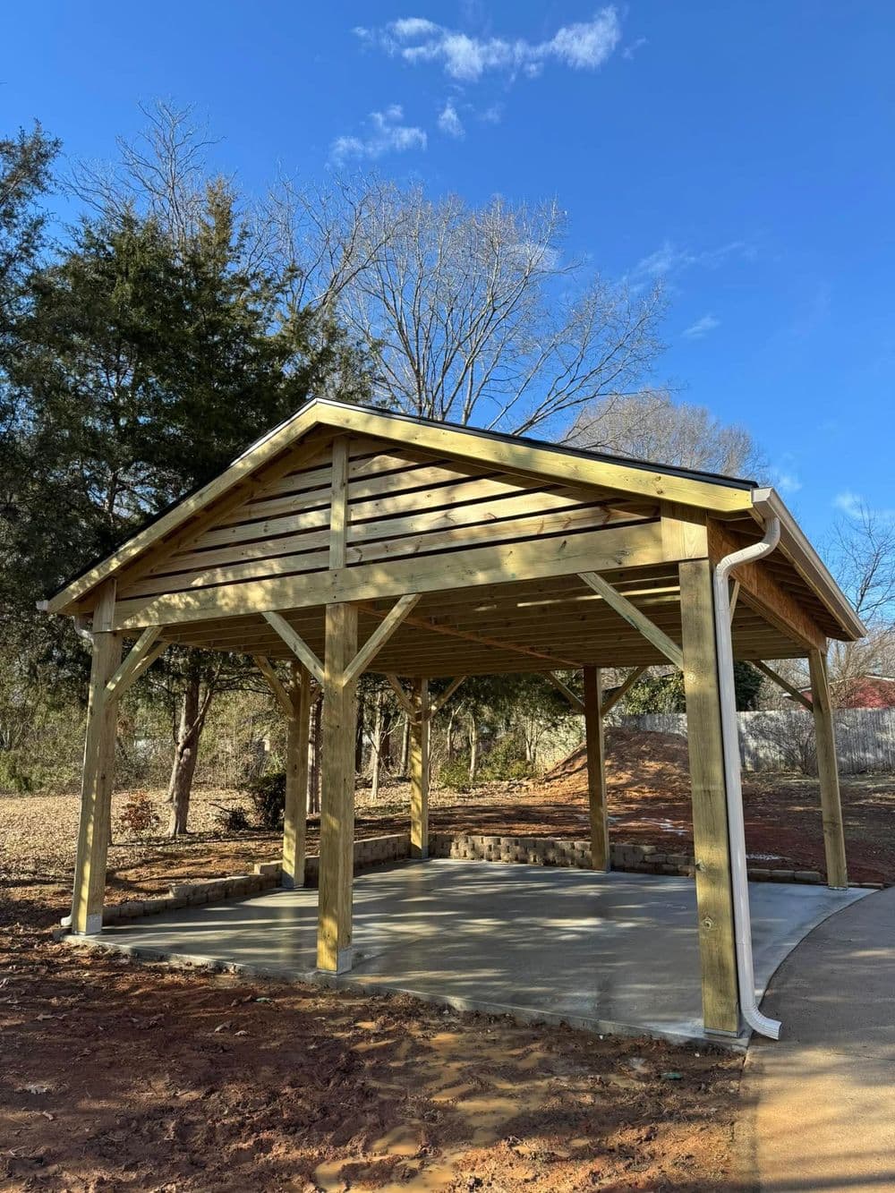 Wooden pavilion with a concrete floor, surrounded by trees and clear blue sky.