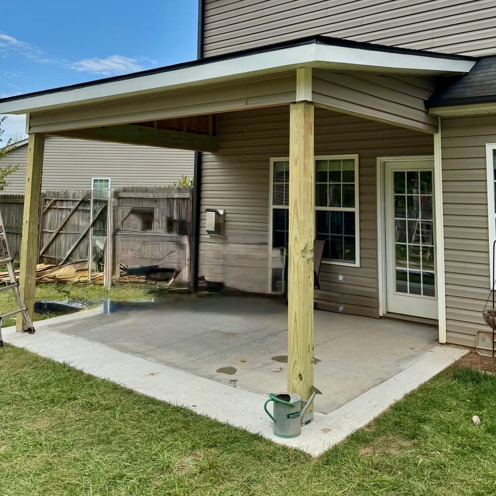 Covered patio area with concrete floor and grass, adjacent to a house and backyard.