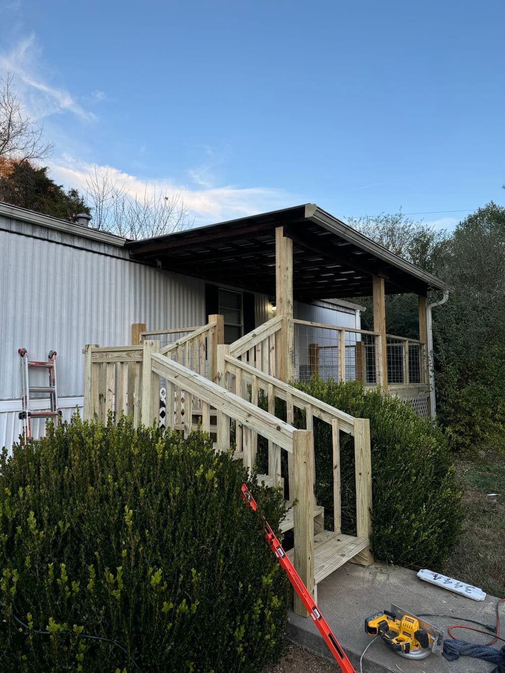 Wooden ramp and porch renovation on a mobile home, surrounded by greenery. Tools visible.