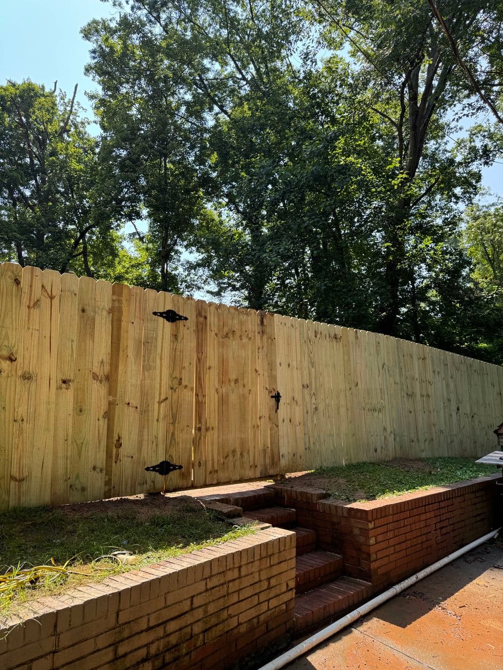 Wooden fence with black hardware, surrounded by lush greenery and brick stairs in backyard.
