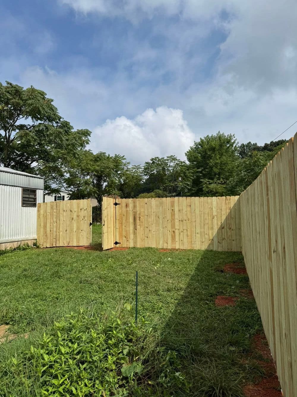 Wooden backyard fence with gate surrounded by grass and trees under a cloudy sky.