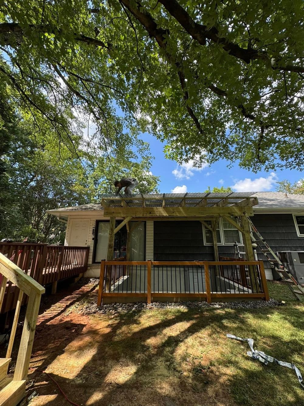 Construction worker installing a wooden deck on a house surrounded by trees under a blue sky.