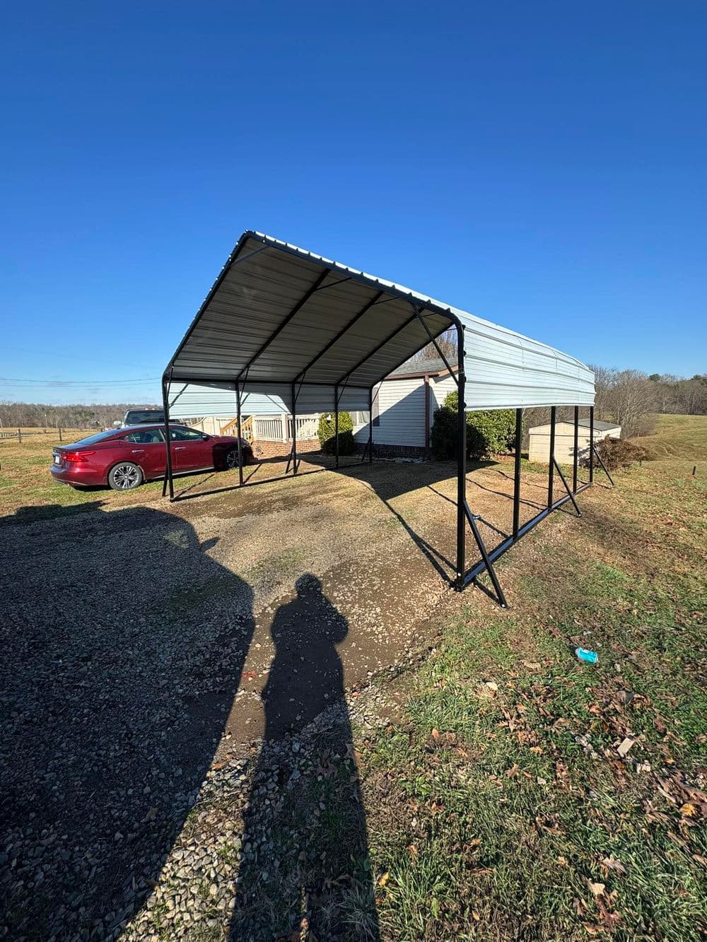 Metal carport structure providing shade for vehicles in a rural setting. Clear blue sky backdrop.