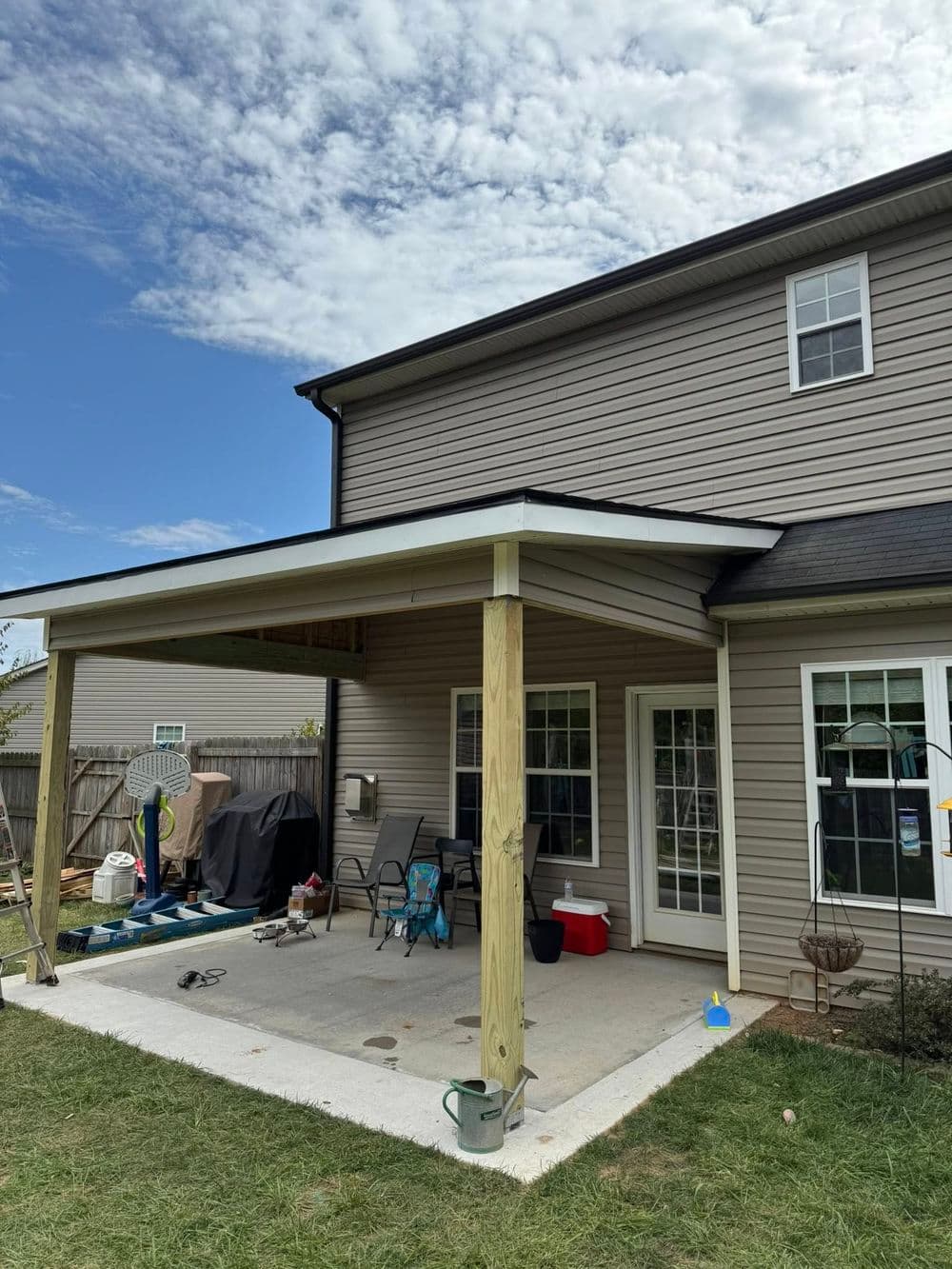 Backyard patio with overhang, chairs, and grill under a blue sky. Perfect for outdoor gatherings.
