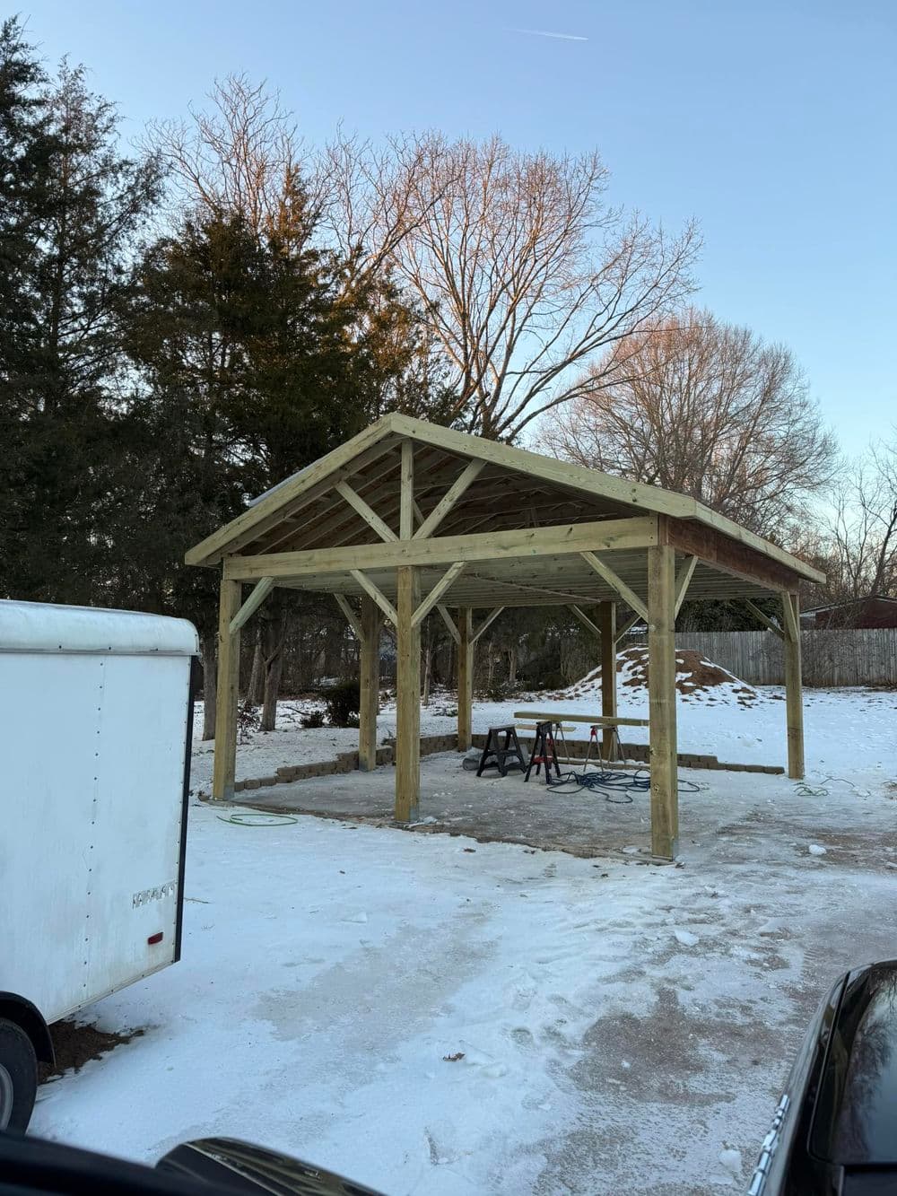 Wooden open pavilion structure in a snowy outdoor setting, surrounded by trees and equipment.